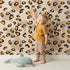 Lovely child standing in front of Whimsy Tots Playful Cub Spots Wallpaper in Beige with leopard, spot, pattern in design. Lovely child standing in front of Whimsy Tots Playful Cub Spots Wallpaper in Beige with leopard, spot, pattern in design.