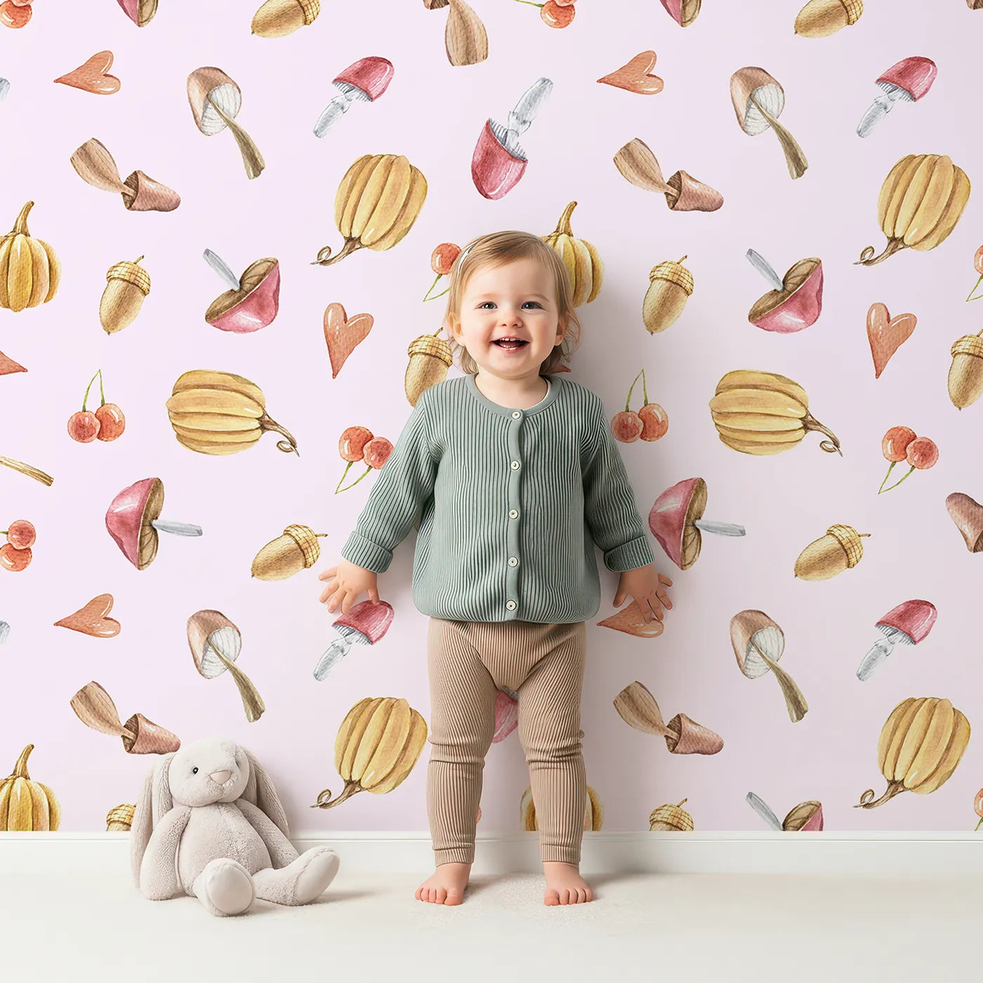 Lovely child standing in front of Whimsy Tots Autumn Forage Friends Wallpaper in Blush Pink with mushroom, acorn, cherry, pumpkin, heart in design.