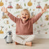 Lovely child standing in front of Whimsy Tots Wishful Woodland Friends Wallpaper in White with hedgehog, owl, mouse, snail, mushroom, leaf, pumpkin, star in design. Lovely child standing in front of Whimsy Tots Wishful Woodland Friends Wallpaper in White with hedgehog, owl, mouse, snail, mushroom, leaf, pumpkin, star in design.