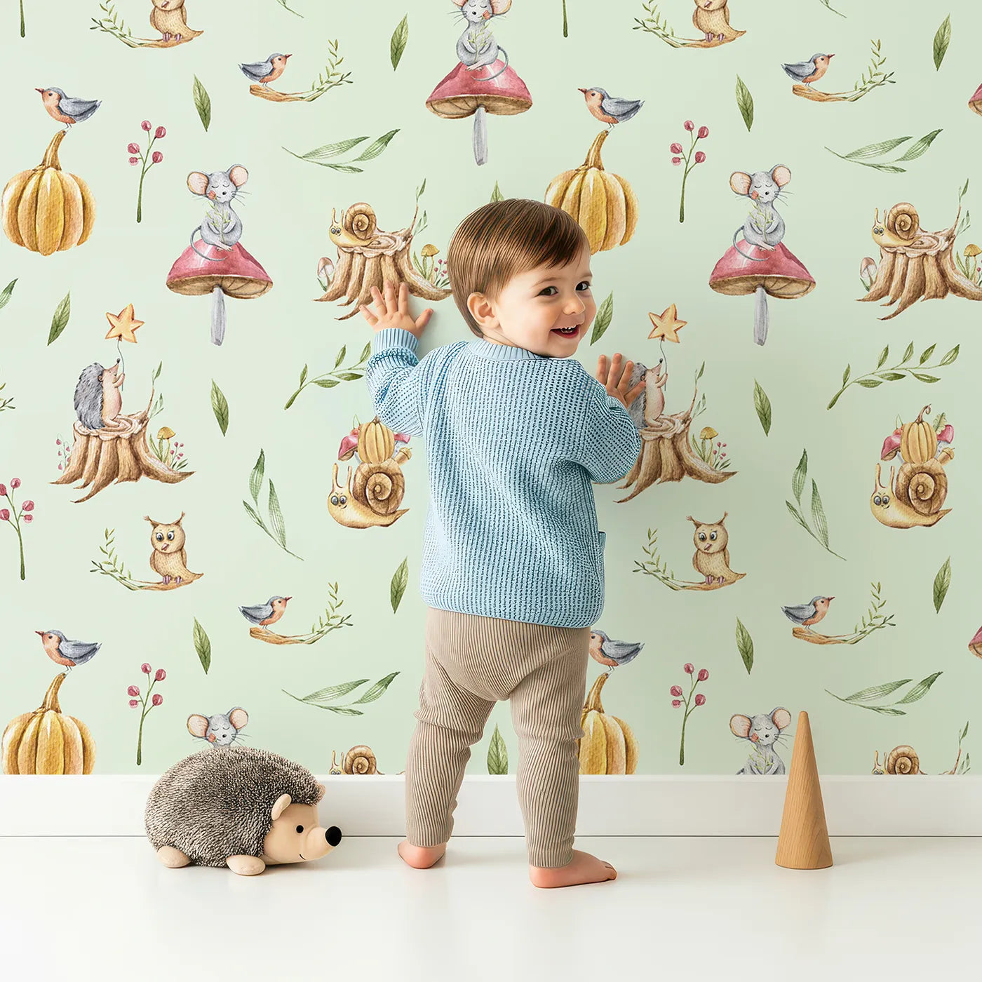 Lovely child standing in front of Whimsy Tots Wishful Woodland Friends Wallpaper in Mint Green with hedgehog, owl, mouse, snail, mushroom, leaf, pumpkin, star in design.