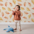 Lovely child standing in front of Whimsy Tots Falling Oak Whispers Wallpaper in Cream with oak leaf, leaf in design. Lovely child standing in front of Whimsy Tots Falling Oak Whispers Wallpaper in Cream with oak leaf, leaf in design.