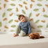 Lovely child standing in front of Whimsy Tots Acorn Meadow Whimsy Wallpaper in White with acorn, oak leaf, leaf in design. Lovely child standing in front of Whimsy Tots Acorn Meadow Whimsy Wallpaper in White with acorn, oak leaf, leaf in design.