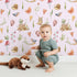 Lovely child standing in front of Whimsy Tots Woodland Playtime Parade Wallpaper in Blush Pink with owl, bird, butterfly, snail, mushroom, heart, stump in design. Lovely child standing in front of Whimsy Tots Woodland Playtime Parade Wallpaper in Blush Pink with owl, bird, butterfly, snail, mushroom, heart, stump in design.