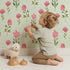 Lovely child standing in front of Whimsy Tots Pink Meadow Blossoms Wallpaper in Mint with flower, leaf in design. Lovely child standing in front of Whimsy Tots Pink Meadow Blossoms Wallpaper in Mint with flower, leaf in design.