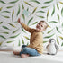 Lovely child standing in front of Whimsy Tots Whimsy Leaf Scatter Wallpaper in White with leaf, spray, greenery, foliage in design. Lovely child standing in front of Whimsy Tots Whimsy Leaf Scatter Wallpaper in White with leaf, spray, greenery, foliage in design.