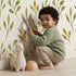 Lovely child standing in front of Whimsy Tots Whimsy Leaf Branch Wallpaper in White with leaf, stem, greenery, branch in design. Lovely child standing in front of Whimsy Tots Whimsy Leaf Branch Wallpaper in White with leaf, stem, greenery, branch in design.