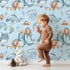 Lovely child standing in front of Whimsy Tots Whale Balloon Parade Wallpaper in Sky Blue with whale, balloon, octopus, bunting, seaweed, crown in design. Lovely child standing in front of Whimsy Tots Whale Balloon Parade Wallpaper in Sky Blue with whale, balloon, octopus, bunting, seaweed, crown in design.