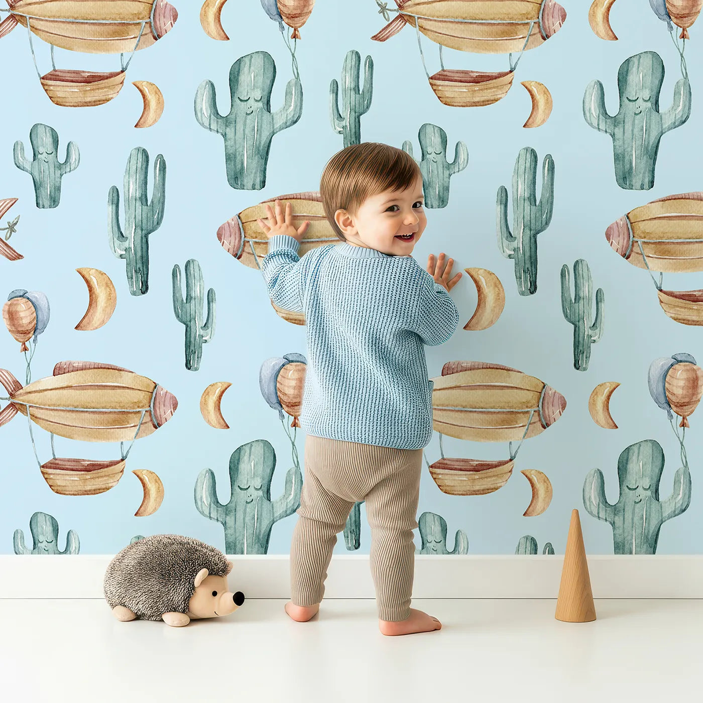 Lovely child standing in front of Whimsy Tots Desert Balloon Voyage Wallpaper in Blue with airship, cactus, moon, balloon in design.