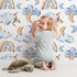 Lovely child standing in front of Whimsy Tots Sleepy Skies Wallpaper in White with cloud, moon, rainbow, sheep in design. Lovely child standing in front of Whimsy Tots Sleepy Skies Wallpaper in White with cloud, moon, rainbow, sheep in design.