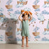 Lovely child standing in front of Whimsy Tots Cloud Sailor Dreams Wallpaper in Blush Pink with ship, cloud, sheep, star, sail in design. Lovely child standing in front of Whimsy Tots Cloud Sailor Dreams Wallpaper in Blush Pink with ship, cloud, sheep, star, sail in design.