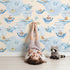 Lovely child standing in front of Whimsy Tots Whale Boat Parade Wallpaper in Cream with whale, submarine, boat, octopus, anchor, flag in design. Lovely child standing in front of Whimsy Tots Whale Boat Parade Wallpaper in Cream with whale, submarine, boat, octopus, anchor, flag in design.