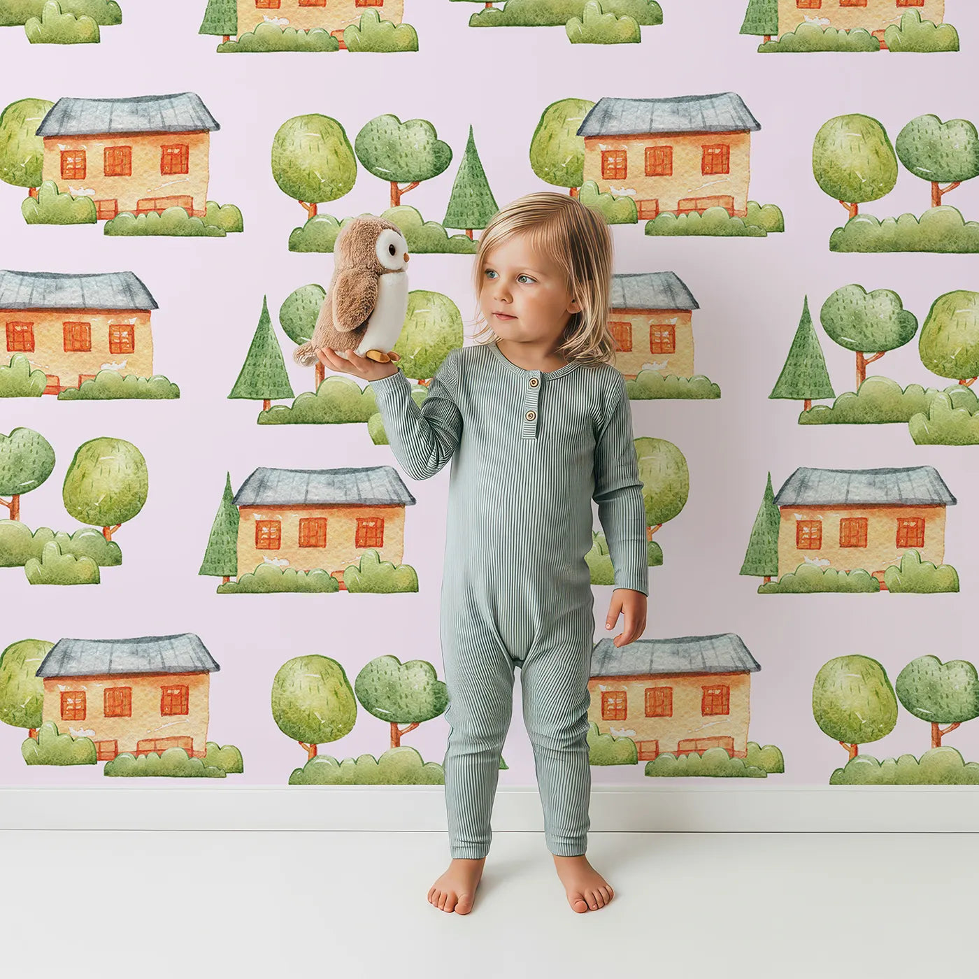 Lovely child standing in front of Whimsy Tots Cottage Garden Village Wallpaper in Pink with tree, bush, house in design.