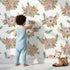 Lovely child standing in front of Whimsy Tots Rustic Blush Bouquet Wallpaper in White with flower, leaf, palm in design. Lovely child standing in front of Whimsy Tots Rustic Blush Bouquet Wallpaper in White with flower, leaf, palm in design.