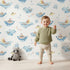 Lovely child standing in front of Whimsy Tots Whale Submarine Adventure Wallpaper in White with whale, submarine, fish, anchor, octopus, bunting in design. Lovely child standing in front of Whimsy Tots Whale Submarine Adventure Wallpaper in White with whale, submarine, fish, anchor, octopus, bunting in design.