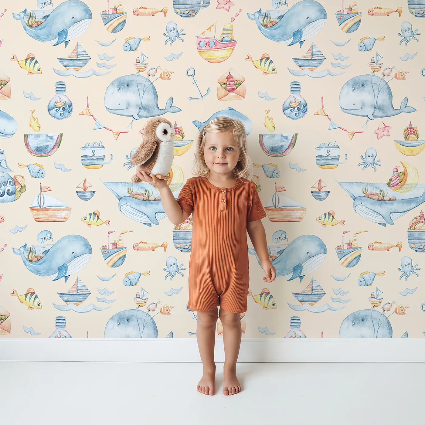 Lovely child standing in front of Whimsy Tots Whale Mail Wallpaper in Peach with whale, fish, octopus, boat, lighthouse, anchor, bird, mug, watermelon, letter in design.