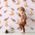 Lovely child standing in front of Whimsy Tots Moon Perch Birds Wallpaper in Pink with bird, moon in design. Lovely child standing in front of Whimsy Tots Moon Perch Birds Wallpaper in Pink with bird, moon in design.