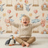 Lovely child standing in front of Whimsy Tots Dreamland Friends Wallpaper in Beige with bird, bunny, cloud, balloon, star in design. Lovely child standing in front of Whimsy Tots Dreamland Friends Wallpaper in Beige with bird, bunny, cloud, balloon, star in design.