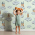Lovely child standing in front of Whimsy Tots Castle Cloud Drift Wallpaper in Mint Green with castle, cloud, moon in design. Lovely child standing in front of Whimsy Tots Castle Cloud Drift Wallpaper in Mint Green with castle, cloud, moon in design.