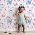 Lovely child standing in front of Whimsy Tots Starlight Narwhal Parade Wallpaper in Pink with narwhal, unicorn, moon, star, cloud, heart, fruit in design. Lovely child standing in front of Whimsy Tots Starlight Narwhal Parade Wallpaper in Pink with narwhal, unicorn, moon, star, cloud, heart, fruit in design.