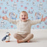 Lovely child standing in front of Whimsy Tots Starlight Narwhal Parade Wallpaper in Blue with narwhal, unicorn, moon, star, cloud, heart, fruit in design. Lovely child standing in front of Whimsy Tots Starlight Narwhal Parade Wallpaper in Blue with narwhal, unicorn, moon, star, cloud, heart, fruit in design.