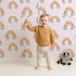 Lovely child standing in front of Whimsy Tots Sunset Rainbows Wallpaper in Pink with rainbow, cloud in design. Lovely child standing in front of Whimsy Tots Sunset Rainbows Wallpaper in Pink with rainbow, cloud in design.
