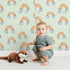 Lovely child standing in front of Whimsy Tots Sunset Rainbows Wallpaper in Green with rainbow, cloud in design. Lovely child standing in front of Whimsy Tots Sunset Rainbows Wallpaper in Green with rainbow, cloud in design.