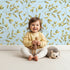 Lovely child standing in front of Whimsy Tots Golden Olive Garden Wallpaper in Sky with leaf, olive, sprig, stem in design. Lovely child standing in front of Whimsy Tots Golden Olive Garden Wallpaper in Sky with leaf, olive, sprig, stem in design.