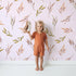 Lovely child standing in front of Whimsy Tots Sunset Meadow Breeze Wallpaper in Blush Pink with leaf, stem, sprig in design. Lovely child standing in front of Whimsy Tots Sunset Meadow Breeze Wallpaper in Blush Pink with leaf, stem, sprig in design.