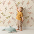 Lovely child standing in front of Whimsy Tots Sunset Meadow Breeze Wallpaper in Cream with leaf, stem, sprig, branch in design. Lovely child standing in front of Whimsy Tots Sunset Meadow Breeze Wallpaper in Cream with leaf, stem, sprig, branch in design.