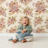 Lovely child standing in front of Whimsy Tots Autumn Bloom Harmony Wallpaper in Cream with flower, leaf, peony, bouquet in design. Lovely child standing in front of Whimsy Tots Autumn Bloom Harmony Wallpaper in Cream with flower, leaf, peony, bouquet in design.