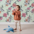 Lovely child standing in front of Whimsy Tots Winter Blooming Garden Wallpaper in Mint Green with flower, cotton, eucalyptus, poinsettia in design. Lovely child standing in front of Whimsy Tots Winter Blooming Garden Wallpaper in Mint Green with flower, cotton, eucalyptus, poinsettia in design.