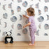 Lovely child standing in front of Whimsy Tots Celestial Winter Blooms Wallpaper in White with moon, flower, cotton, eucalyptus in design. Lovely child standing in front of Whimsy Tots Celestial Winter Blooms Wallpaper in White with moon, flower, cotton, eucalyptus in design.