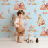 Lovely child standing in front of Whimsy Tots Feathered Serenity Wallpaper in Sky with bird, swan, crown, mountain, wing in design. Lovely child standing in front of Whimsy Tots Feathered Serenity Wallpaper in Sky with bird, swan, crown, mountain, wing in design.