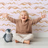 Lovely child standing in front of Whimsy Tots Soft Wave Rhythm Wallpaper in Blush Pink with wave, curve, line, brushstroke, abstract in design. Lovely child standing in front of Whimsy Tots Soft Wave Rhythm Wallpaper in Blush Pink with wave, curve, line, brushstroke, abstract in design.