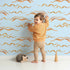 Lovely child standing in front of Whimsy Tots Soft Wave Rhythm Wallpaper in Sky Blue with wave, curve, line, brushstroke, abstract in design. Lovely child standing in front of Whimsy Tots Soft Wave Rhythm Wallpaper in Sky Blue with wave, curve, line, brushstroke, abstract in design.
