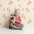 Lovely child standing in front of Whimsy Tots Rustling Meadow Sprigs Wallpaper in Sand with leaf, sprig, botanical, plant in design. Lovely child standing in front of Whimsy Tots Rustling Meadow Sprigs Wallpaper in Sand with leaf, sprig, botanical, plant in design.