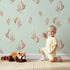 Lovely child standing in front of Whimsy Tots Rustling Meadow Sprigs Wallpaper in Mint Green with leaf, sprig, botanical, plant in design. Lovely child standing in front of Whimsy Tots Rustling Meadow Sprigs Wallpaper in Mint Green with leaf, sprig, botanical, plant in design.