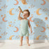 Lovely child standing in front of Whimsy Tots Sleepy Skies Shine Wallpaper in Blue with moon, star, sky, sleepy face in design. Lovely child standing in front of Whimsy Tots Sleepy Skies Shine Wallpaper in Blue with moon, star, sky, sleepy face in design.