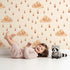 Lovely child standing in front of Whimsy Tots Rainy Day Nap Wallpaper in Cream with cloud, raindrop, sleepy face in design. Lovely child standing in front of Whimsy Tots Rainy Day Nap Wallpaper in Cream with cloud, raindrop, sleepy face in design.
