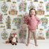 Lovely child standing in front of Whimsy Tots Whimsy Village Dreams Wallpaper in White with house, rainbow, cup, jar, cloud, heart, balloon in design. Lovely child standing in front of Whimsy Tots Whimsy Village Dreams Wallpaper in White with house, rainbow, cup, jar, cloud, heart, balloon in design.