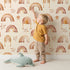 Lovely child standing in front of Whimsy Tots Golden Boho Rainbows Wallpaper in Beige with rainbow, star in design. Lovely child standing in front of Whimsy Tots Golden Boho Rainbows Wallpaper in Beige with rainbow, star in design.