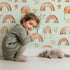 Lovely child standing in front of Whimsy Tots Golden Boho Rainbows Wallpaper in Mint with rainbow, star in design. Lovely child standing in front of Whimsy Tots Golden Boho Rainbows Wallpaper in Mint with rainbow, star in design.