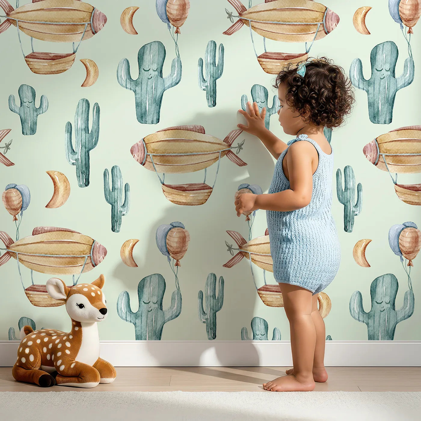 Lovely child standing in front of Whimsy Tots Cactus Cloud Voyage Wallpaper in Sage Green with cactus, airship, balloon, moon in design.