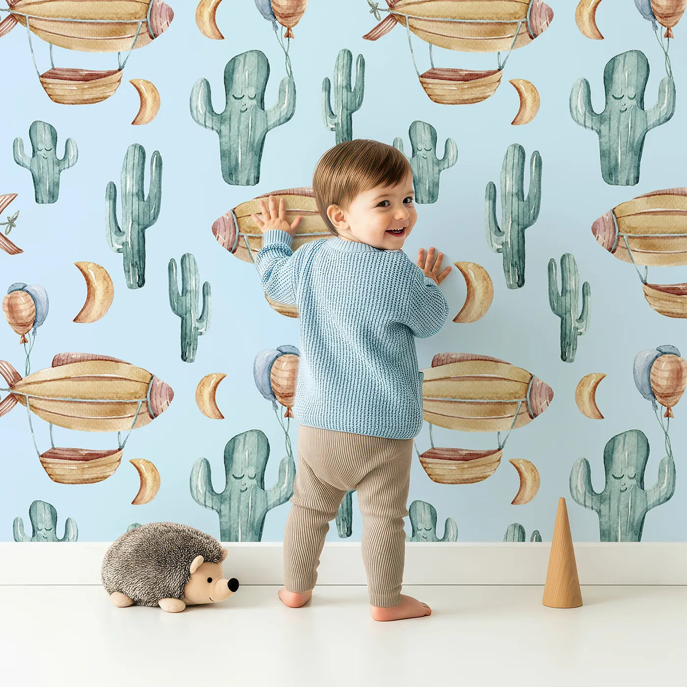Lovely child standing in front of Whimsy Tots Cactus Cloud Voyage Wallpaper in Sky Blue with cactus, airship, balloon, moon in design.