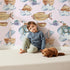 Lovely child standing in front of Whimsy Tots Skybound Explorers Wallpaper in Blush Pink with submarine, boat, fish, cloud, airship in design. Lovely child standing in front of Whimsy Tots Skybound Explorers Wallpaper in Blush Pink with submarine, boat, fish, cloud, airship in design.