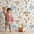 Lovely child standing in front of Whimsy Tots Sky Dreams Celebration Wallpaper in White with airship, balloon, crown, moon, rainbow, heart, elephant, bird in design. Lovely child standing in front of Whimsy Tots Sky Dreams Celebration Wallpaper in White with airship, balloon, crown, moon, rainbow, heart, elephant, bird in design.