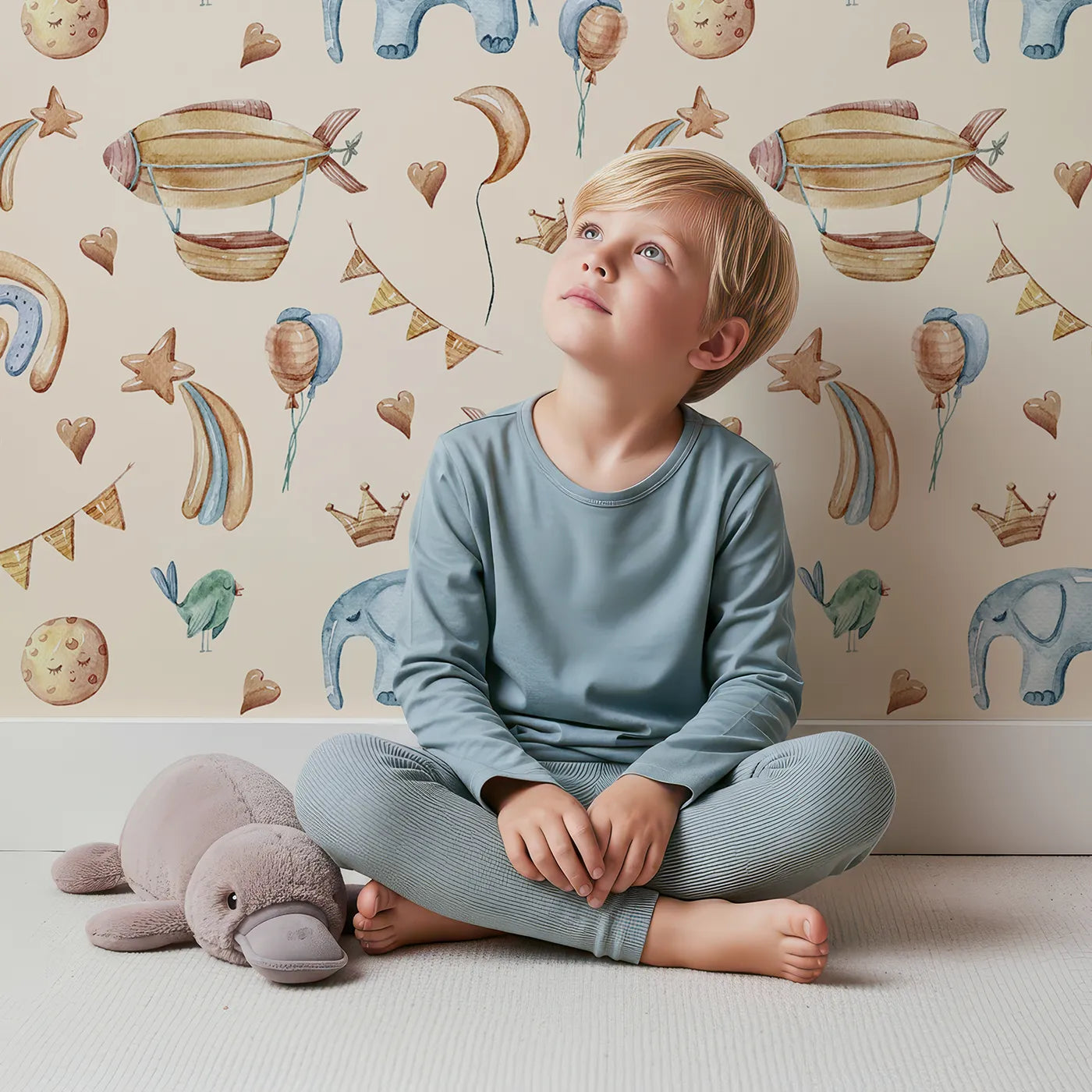 Lovely child standing in front of Whimsy Tots Sky Dreams Celebration Wallpaper in Beige with airship, balloon, crown, moon, rainbow, heart, elephant, bird in design.