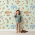 Lovely child standing in front of Whimsy Tots Sky Dreams Celebration Wallpaper in Mint Green with airship, balloon, crown, moon, rainbow, heart, elephant, bird in design. Lovely child standing in front of Whimsy Tots Sky Dreams Celebration Wallpaper in Mint Green with airship, balloon, crown, moon, rainbow, heart, elephant, bird in design.