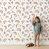 Lovely child standing in front of Whimsy Tots Sky Bloom Party Wallpaper in White with rainbow, balloon, moon, star, bunting, heart in design. Lovely child standing in front of Whimsy Tots Sky Bloom Party Wallpaper in White with rainbow, balloon, moon, star, bunting, heart in design.