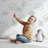 Lovely child standing in front of Whimsy Tots Petal Bloom Grace Wallpaper in White with flower, leaf, sprig, blossom in design. Lovely child standing in front of Whimsy Tots Petal Bloom Grace Wallpaper in White with flower, leaf, sprig, blossom in design.